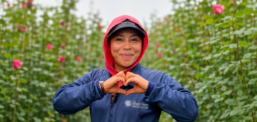 Person in a blue jacket and red hood forms a heart shape with their hands in a rose garden.